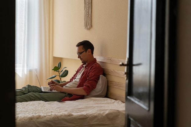 Un jeune homme de race blanche concentré travaille comme journaliste Internet en tapant un article assis sur le lit de la maison
