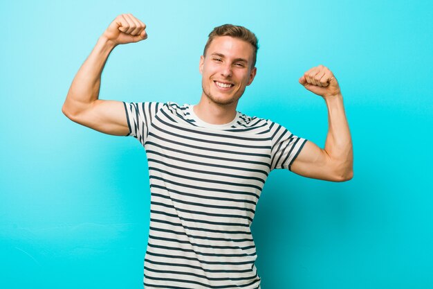 Jeune homme de race blanche contre un mur bleu montrant le geste de la force avec les bras, symbole du pouvoir féminin