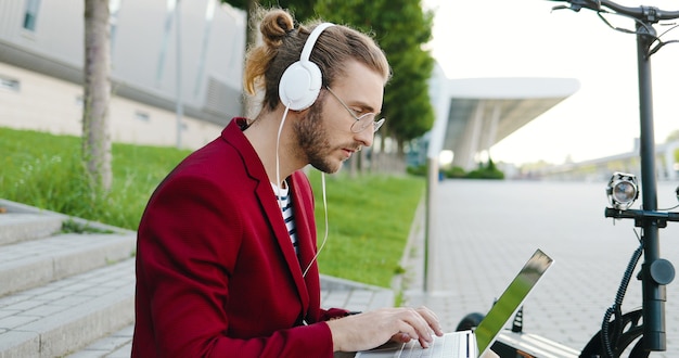 Jeune homme de race blanche dans des lunettes et des écouteurs assis sur les marches en plein air, travaillant sur un ordinateur portable, en tapant sur le clavier. Un gars élégant travaille dans la rue en tant que pigiste. Scooter électrique. Écouter de la musique.