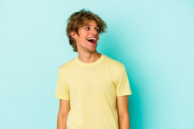 Jeune homme de race blanche avec du maquillage isolé sur fond bleu riant détendu et heureux, cou tendu montrant les dents.