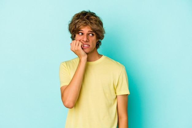 Jeune homme de race blanche avec du maquillage isolé sur fond bleu se rongeant les ongles, nerveux et très anxieux.