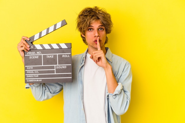 Jeune homme de race blanche avec du maquillage tenant un clap isolé sur fond jaune gardant un secret ou demandant le silence.