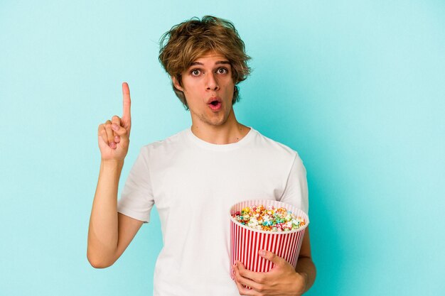 Jeune homme de race blanche avec du maquillage tenant du pop-corn isolé sur fond bleu ayant une bonne idée, concept de créativité.