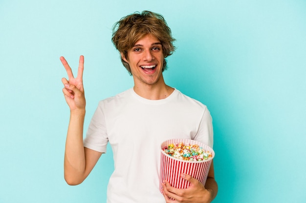 Jeune homme de race blanche avec du maquillage tenant du pop-corn isolé sur fond bleu joyeux et insouciant montrant un symbole de paix avec les doigts.