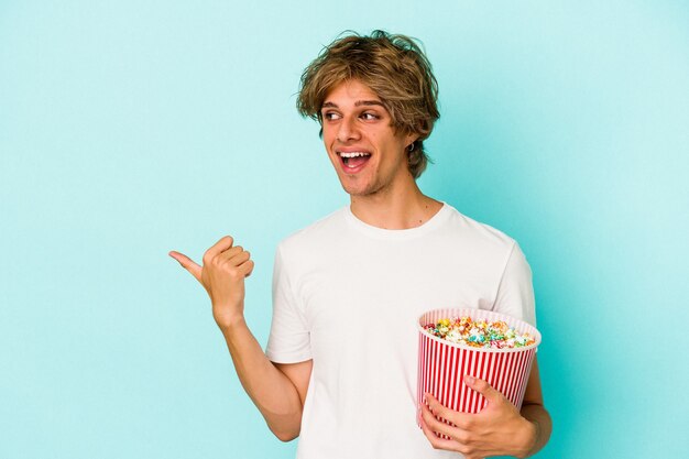 Jeune homme de race blanche avec du maquillage tenant du pop-corn isolé sur des points de fond bleu avec le pouce loin, riant et insouciant.