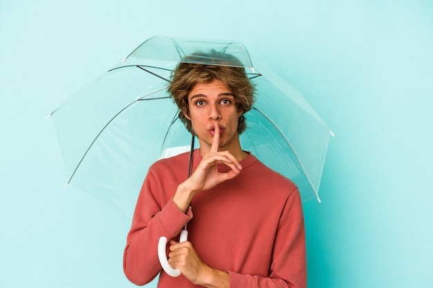 Jeune homme de race blanche avec du maquillage tenant un parapluie isolé sur fond bleu gardant un secret ou demandant le silence.