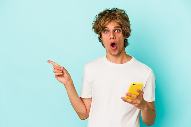 Jeune homme de race blanche avec du maquillage tenant un téléphone portable isolé sur fond bleu pointant vers le côté