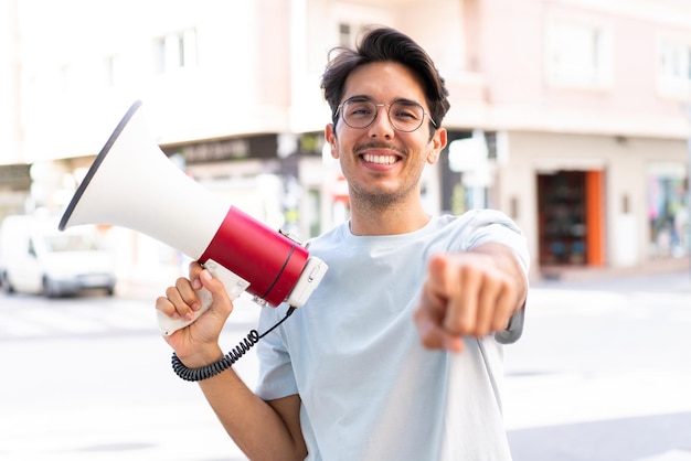 Jeune homme de race blanche à l'extérieur tenant un mégaphone et souriant tout en pointant vers l'avant