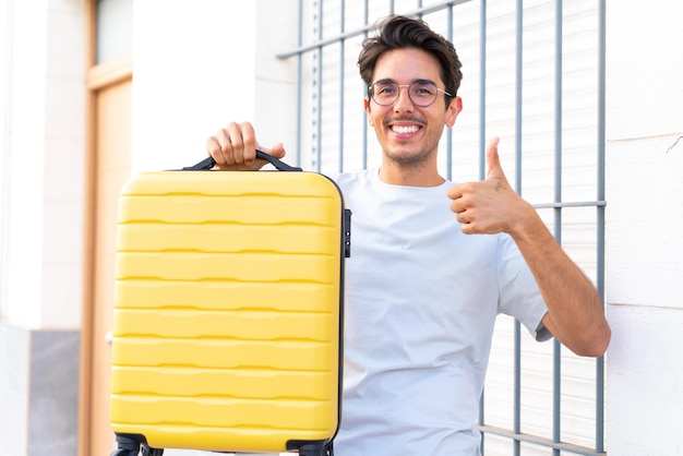 Jeune homme de race blanche à l'extérieur en vacances avec valise de voyage et avec le pouce vers le haut