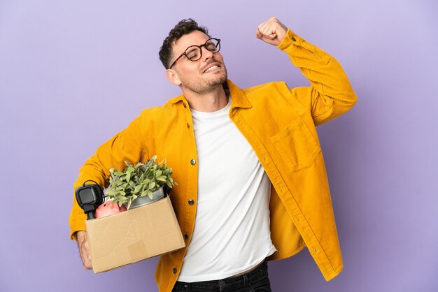 Jeune homme de race blanche faire un mouvement tout en ramassant une boîte pleine de choses isolées sur un mur violet célébrant une victoire