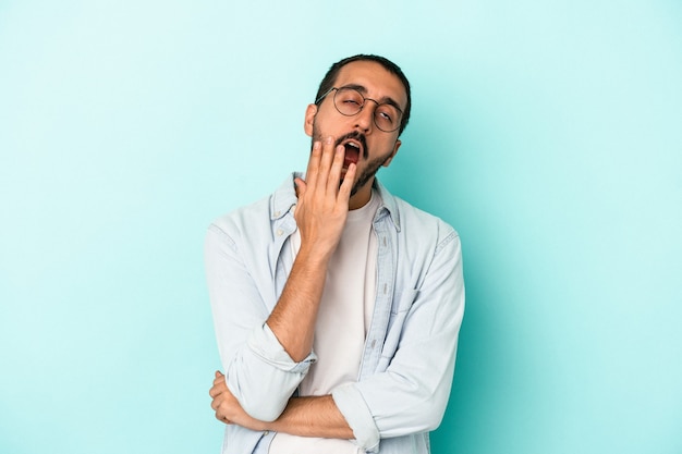 Jeune homme de race blanche isolé sur fond bleu bâillant montrant un geste fatigué couvrant la bouche avec la main.