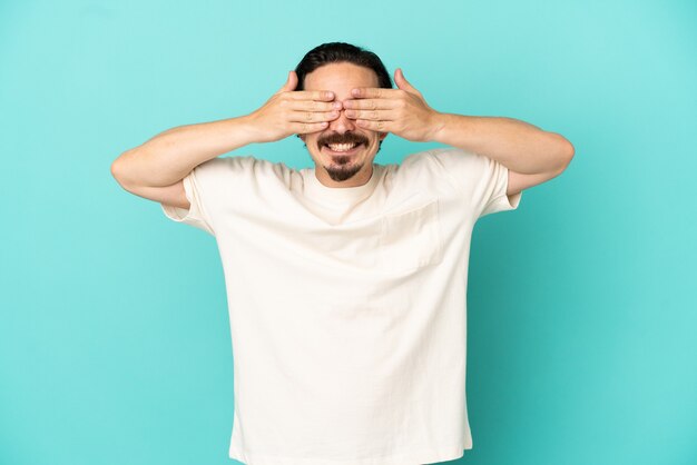 Jeune homme de race blanche isolé sur fond bleu couvrant les yeux par les mains et souriant