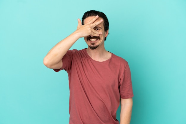 Jeune Homme De Race Blanche Isolé Sur Fond Bleu Couvrant Les Yeux Par Les Mains Et Souriant