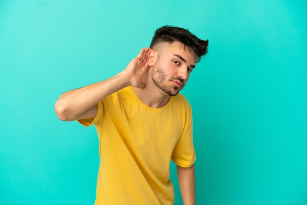 Jeune homme de race blanche isolé sur fond bleu écoutant quelque chose en mettant la main sur l'oreille