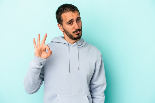 Jeune homme de race blanche isolé sur fond bleu fait un clin d'œil et tient un geste correct avec la main.