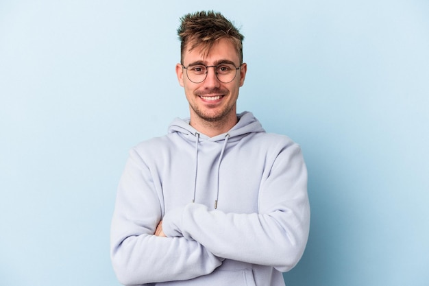 Jeune homme de race blanche isolé sur fond bleu qui se sent confiant, croisant les bras avec détermination.