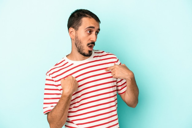 Jeune homme de race blanche isolé sur fond bleu surpris en pointant avec le doigt, souriant largement.