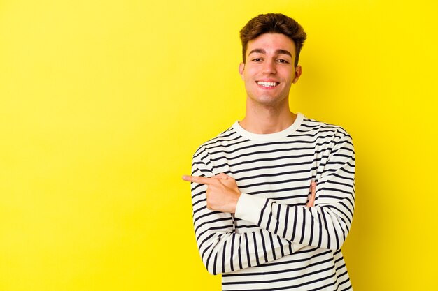 Jeune homme de race blanche isolé sur fond jaune souriant joyeusement pointant avec l'index loin.