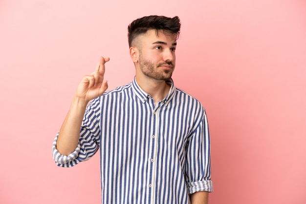 Jeune homme de race blanche isolé sur fond rose avec les doigts croisés et souhaitant le meilleur