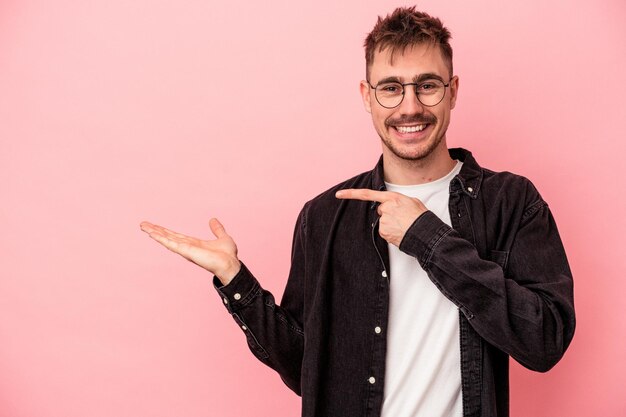 Jeune homme de race blanche isolé sur fond rose excité tenant un espace de copie sur la paume.