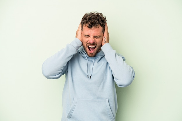 Jeune homme de race blanche isolé sur fond vert couvrant les oreilles avec les mains essayant de ne pas entendre un son trop fort.