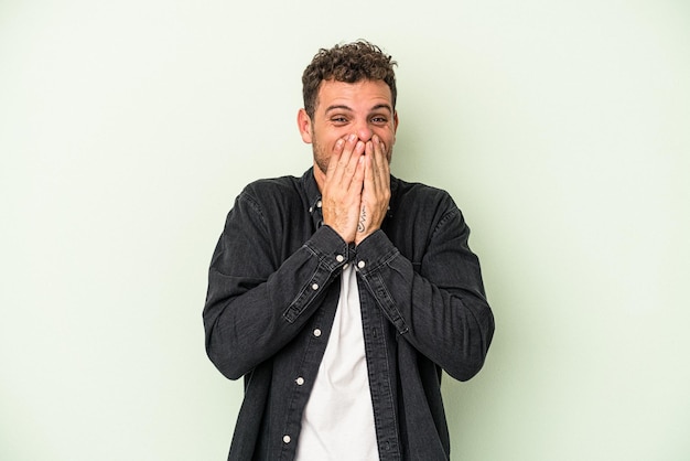 Jeune homme de race blanche isolé sur fond vert en riant de quelque chose, couvrant la bouche avec les mains.