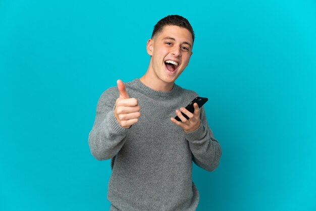 Jeune homme de race blanche isolé sur un mur bleu à l'aide de téléphone mobile tout en faisant les pouces vers le haut