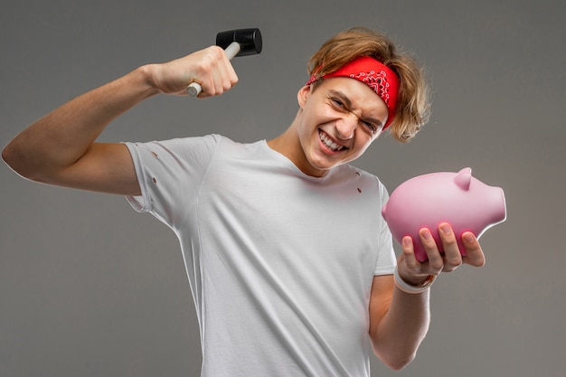 Jeune homme de race blanche à lunettes de soleil rouges, t-shirt blanc avec tirelire cochon rose sur mur gris