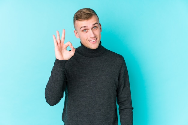 Jeune homme de race blanche sur un mur bleu gai et confiant montrant le geste ok.