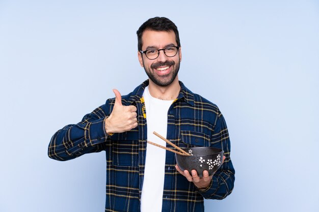 Jeune homme de race blanche sur le mur bleu avec les pouces vers le haut parce que quelque chose de bien s'est produit tout en tenant un bol de nouilles avec des baguettes