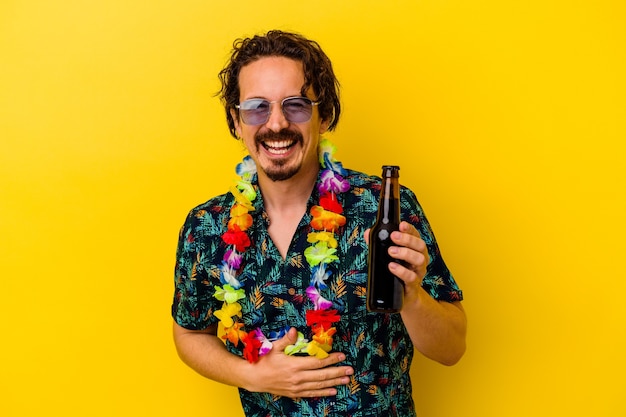 Jeune homme de race blanche portant un collier hawaïen tenant une bière isolée sur un mur jaune en riant et en s'amusant.