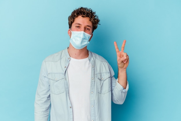 Jeune homme de race blanche portant un masque antiviral isolé sur mur bleu joyeux et insouciant montrant un symbole de paix avec les doigts