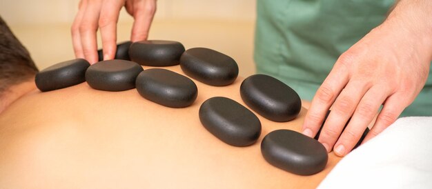 Jeune homme de race blanche recevant un massage du dos avec des pierres noires par un masseur dans un salon de spa. Une femme recevant un soin au spa.