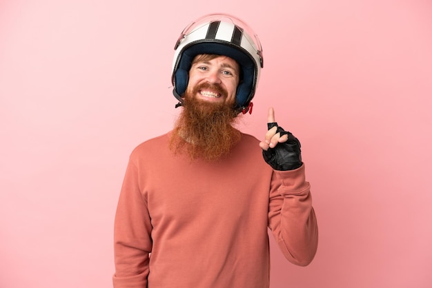 Jeune homme de race blanche rougeâtre avec un casque de moto isolé sur fond rose montrant et levant un doigt en signe du meilleur