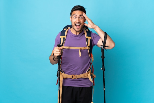 Jeune homme de race blanche avec sac à dos et bâtons de randonnée isolé sur fond bleu avec une expression de surprise