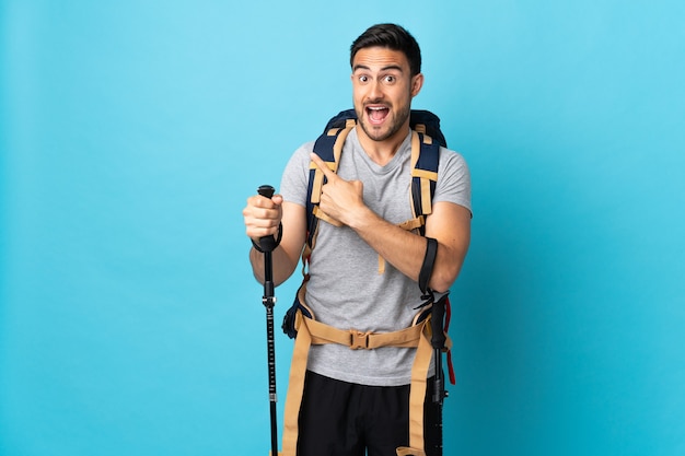 Jeune homme de race blanche avec sac à dos et bâtons de randonnée isolé sur fond bleu surpris et côté pointant