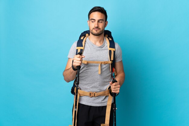 Jeune homme de race blanche avec sac à dos et bâtons de randonnée isolés sur mur bleu faisant des doutes à côté de geste