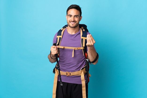 Jeune homme de race blanche avec sac à dos et bâtons de randonnée isolés sur mur bleu faisant le geste de l'argent