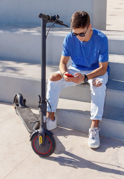 Jeune homme de race blanche avec smartphone assis en plein air près de scooter électrique et à l'aide de smartphone