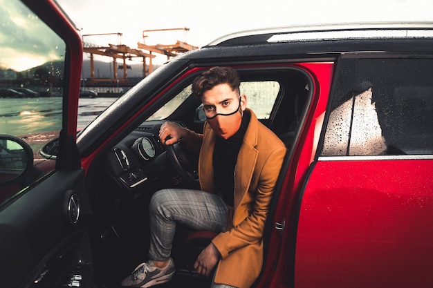 Jeune homme de race blanche sortant d'une voiture rouge portant un masque sur un parking.