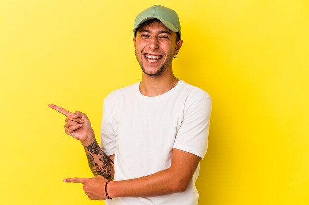 Jeune homme de race blanche avec des tatouages isolés sur fond jaune pointant avec les index vers un espace de copie, exprimant l'excitation et le désir.