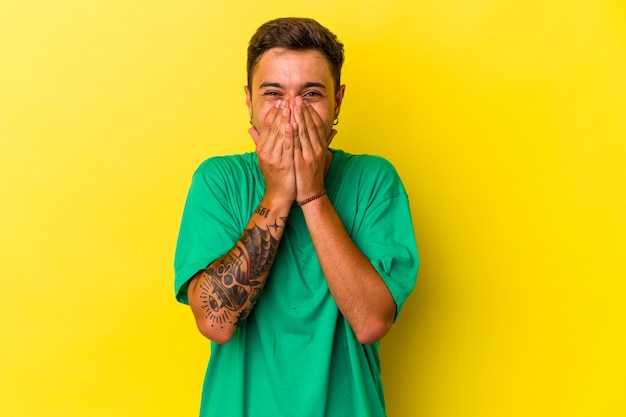 Jeune homme de race blanche avec des tatouages isolés sur fond jaune riant de quelque chose, couvrant la bouche avec les mains.