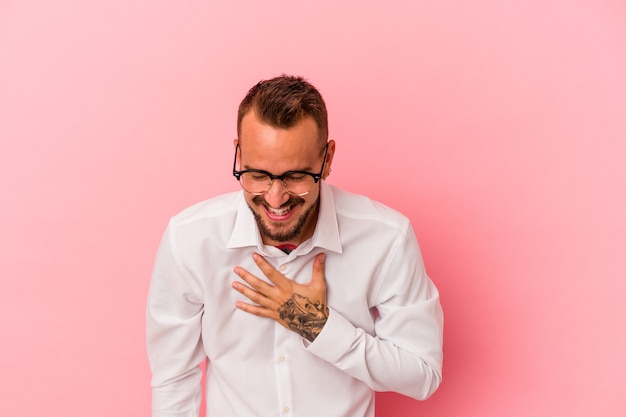 Jeune homme de race blanche avec des tatouages isolés sur fond rose éclate de rire en gardant la main sur la poitrine.