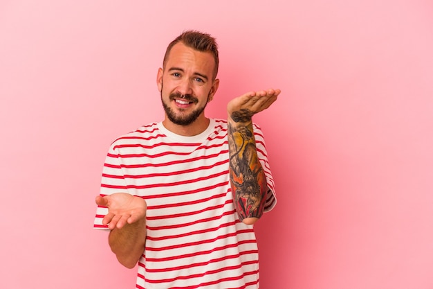 Jeune homme de race blanche avec des tatouages isolés sur fond rose fait l'échelle avec les bras, se sent heureux et confiant.