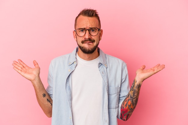 Photo jeune homme de race blanche avec des tatouages isolés sur fond rose haussant les épaules confus et douteux pour tenir un espace de copie.