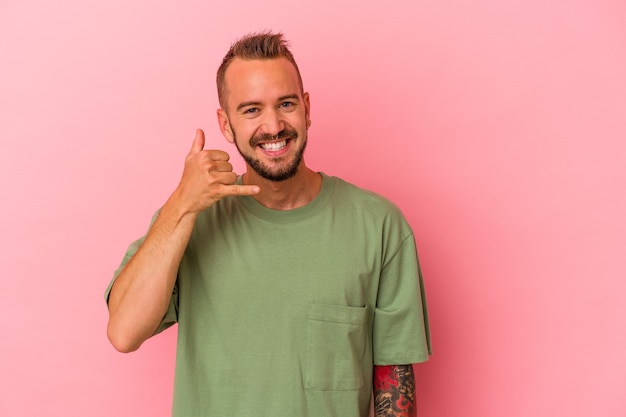 Jeune homme de race blanche avec des tatouages isolés sur fond rose montrant un geste d'appel de téléphone portable avec les doigts.