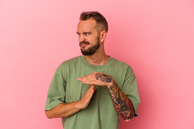 Jeune homme de race blanche avec des tatouages isolés sur fond rose montrant un geste de temporisation.