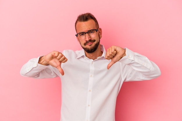 Jeune homme de race blanche avec des tatouages isolés sur fond rose montrant le pouce vers le bas, concept de déception.