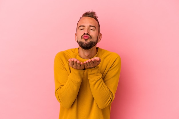 Jeune homme de race blanche avec des tatouages isolés sur fond rose pliant les lèvres et tenant les paumes pour envoyer un baiser aérien.