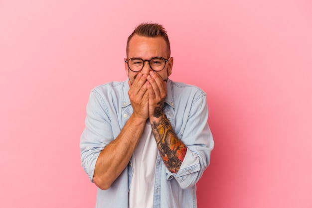 Jeune homme de race blanche avec des tatouages isolés sur fond rose en riant de quelque chose, couvrant la bouche avec les mains.